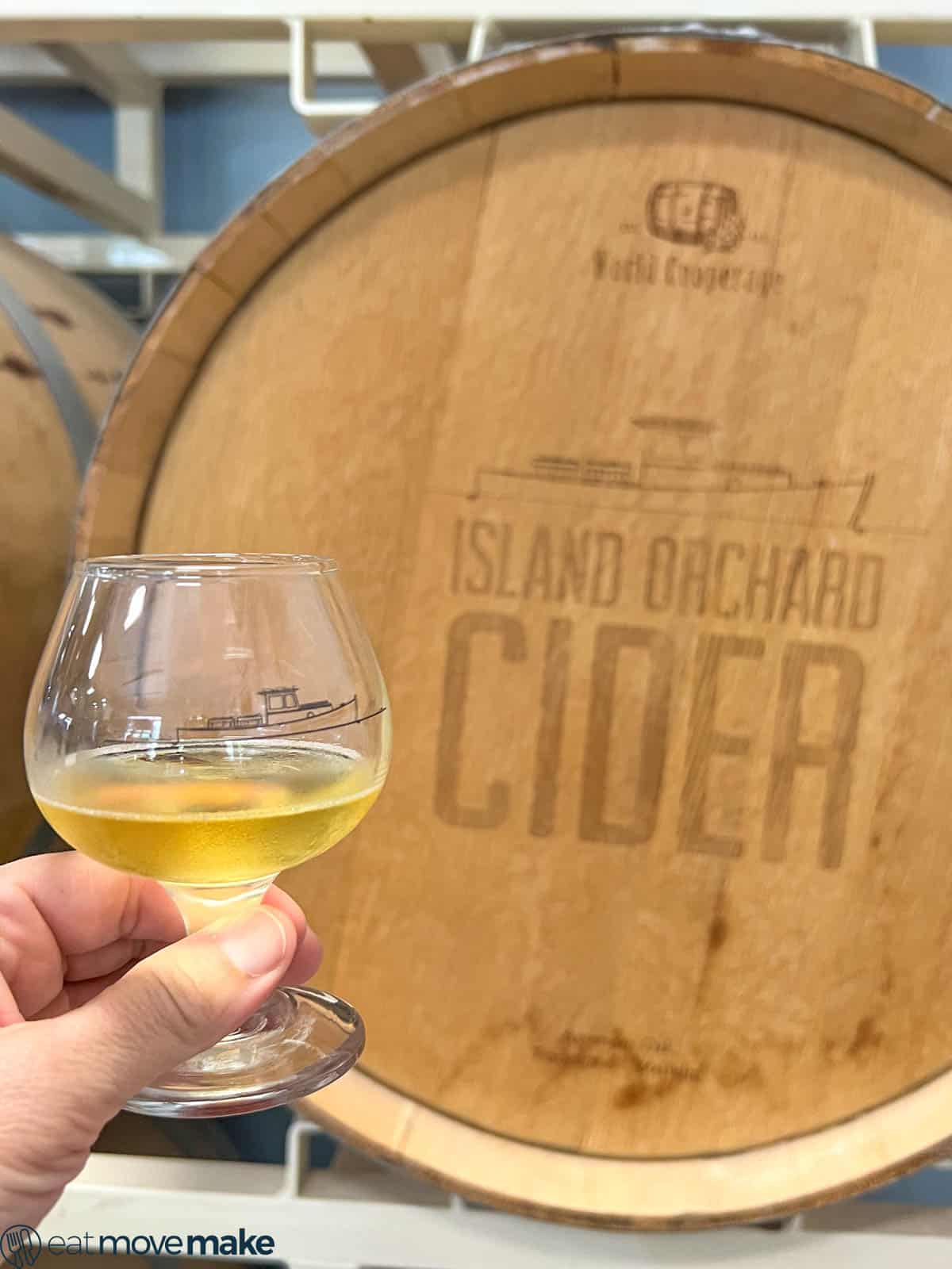 hand holding glass of hard cider by cider barrel