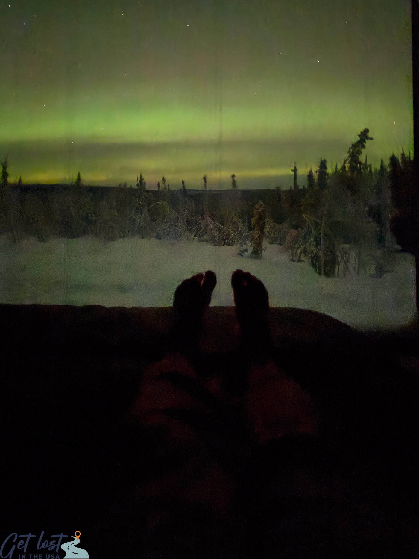 photo of Northern Lights from cube suite at Borealis Basecamp with feet in bed in foreground.